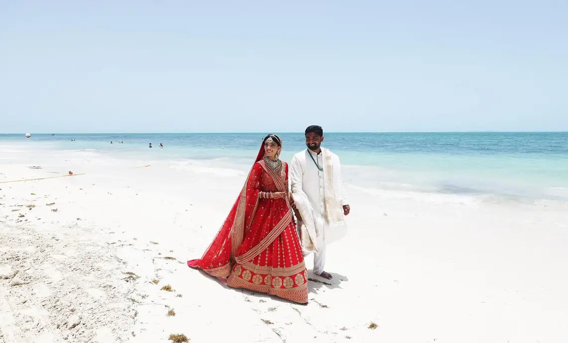 Two people adorned in vibrant South Asian wedding attire stroll barefoot along a pristine, white-sand beach as turquoise waves softly embrace the shore. Under a vast, cloudless blue sky, their joyous presence celebrates love and unity in a setting where cultural tradition meets the beauty of nature from Riviera Maya