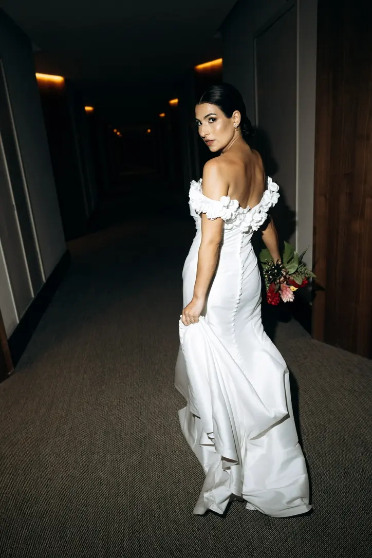 A radiant bride in an off-the-shoulder white gown, bouquet in hand, glances back with a joyful smile as she stands thoughtfully in a softly lit hallway, embodying beauty and love on her special day at grand palladium costa mujeres, cancun