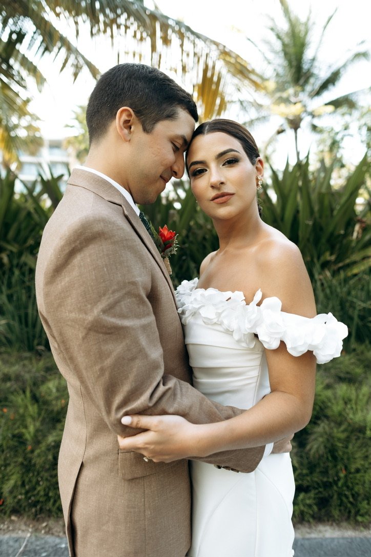 Two people in wedding attire share a tender embrace outdoors, celebrating love in all its forms. One wears a tan suit and gently leans their forehead against their partner, who dazzles in a white dress adorned with delicate floral designs. They&rsquo;re surrounded by vibrant greenery and palm trees swaying in the breeze, creating an enchanting backdrop for this joyful moment.