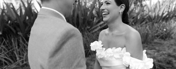 Two partners stand together outdoors, beaming at each other with pure happiness. One wears an elegant off-shoulder dress adorned with vibrant floral designs, while the other sports a suave light-colored suit. Behind them, a tapestry of lush greenery creates a peaceful and welcoming backdrop at grand palladium costa mujeres