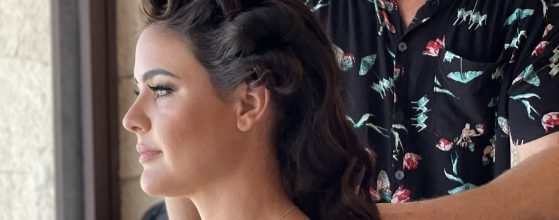 A vibrant woman with beautifully curled hair sits comfortably as Alex corbanezi, hair stylist, wearing a chic black floral shirt, works their magic on her hair. She radiates confidence in her off-shoulder green and white dress, while behind her, a textured beige wall adds warmth and dimension to the inclusive scene.