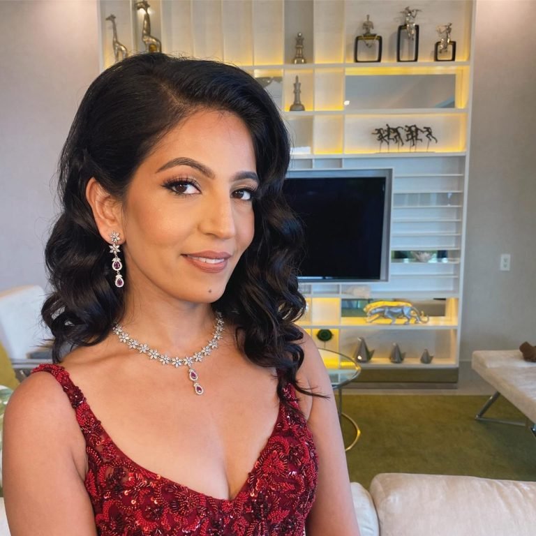 A radiant woman with wavy dark hair shines in a red sequined dress and matching jewelry, her joyful smile lighting up the stylish living room behind her. The space features modern decor, a TV, and shelves filled with character, reflecting both comfort and individuality.