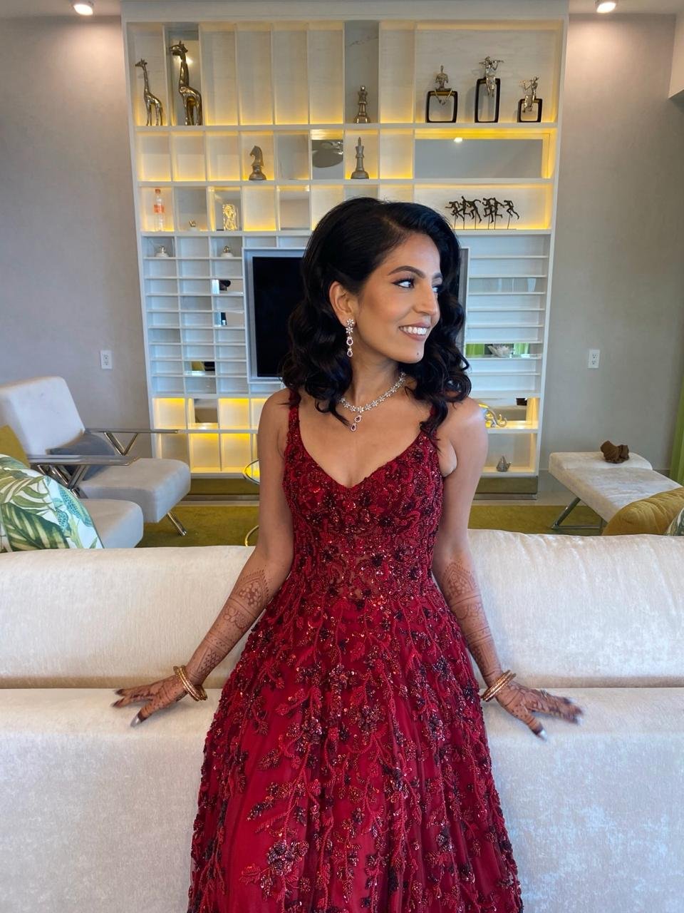 A joyful woman wearing a vibrant red embroidered gown proudly shows off intricate henna designs on her arms as she stands by a white couch, her smile warm and inviting while gazing to the side. Behind her, an illuminated modern shelf displays eclectic décor alongside a TV, creating an atmosphere that celebrates both tradition and contemporary style.